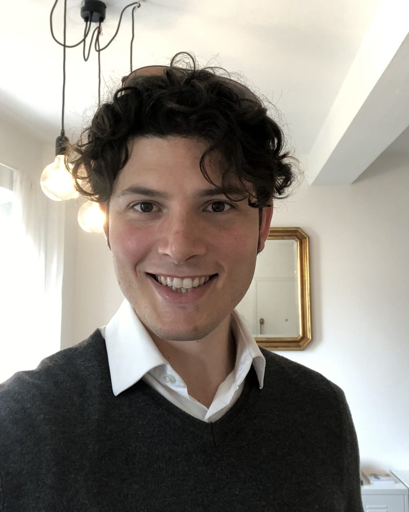 Emanuele Papi, fondatore di Gesturis, sorridente in camicia bianca e maglione grigio in un appartamento a Firenze